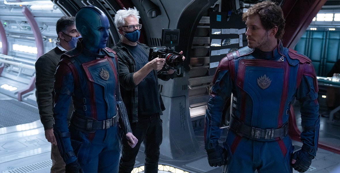 In a behind-the-scenes image from Marvel Studios’ Guardians of the Galaxy Vol. 3, director James Gunn (in blue mask, with white spiky hair, holding a camera) directs Karen Gillan as Nebula (left) and Chris Pratt as Peter Quill (right) on set. Another crew member stands behind and to the left of Gunn. 