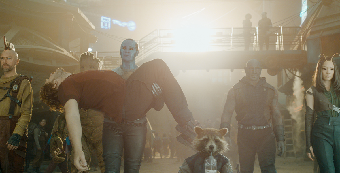 In a scene from Marvel Studios’ Guardians of the Galaxy Vol. 3, from left to right, Kraglin (Sean Gunn), Groot (voiced by Vin Diesel), Nebula (Karen Gillan) holding Peter (Chris Pratt), Rocket (voiced by Bradley Cooper), Drax (Dave Bautista), and Mantis (Pom Klementieff) are walking intently toward the camera.