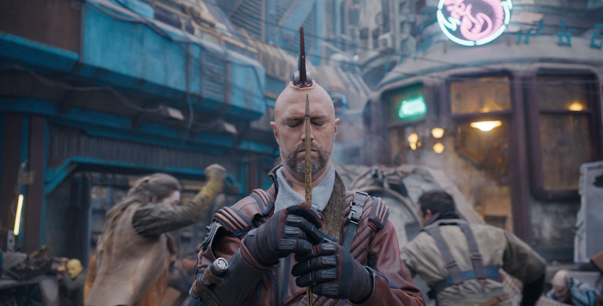 In a still from Marvel Studios’ Guardians of the Galaxy Vol. 3, Kraglin (Sean Gunn) is holding an arrow between his hands, with his eyes closed. There is fighting going on behind him. He is bald, aside from his headpiece-mohawk, and he’s wearing a dark red leather jacket and black and red gloves.