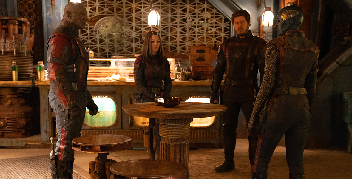 In a still from Marvel Studios’ Guardians of the Galaxy Vol. 3, from left to right, Drax (Dave Bautista), Mantis (Pom Klementieff), Peter Quill (Chris Pratt), and Nebula (Karen Gillan) are standing around a table in a kitchen-like space having a discussion.