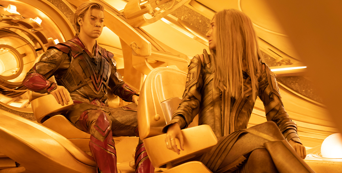 In a still from Marvel Studios’ Guardians of the Galaxy Vol. 3, Adam Warlock (Will Poulter) (top) and Ayesha (Elizabeth Debicki) (bottom) are sitting in golden chairs, in a golden-hued room. Ayesha is turned around, looking at Adam; Adam is staring at something off-camera to the right. They’re both wearing intricate leather costumes. 