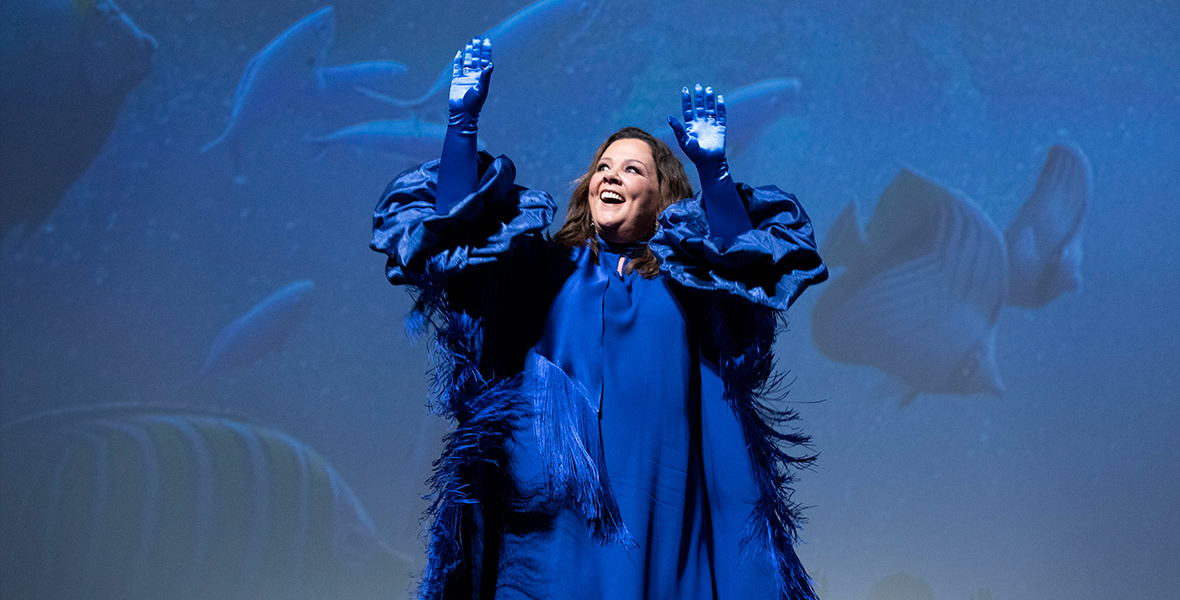 Melissa McCarthy waves to the audience onstage at the Dolby Theater prior to the world premiere screening of The Little Mermaid. She is wearing a deep blue dress embellished with fringe, along with matching blue gloves.