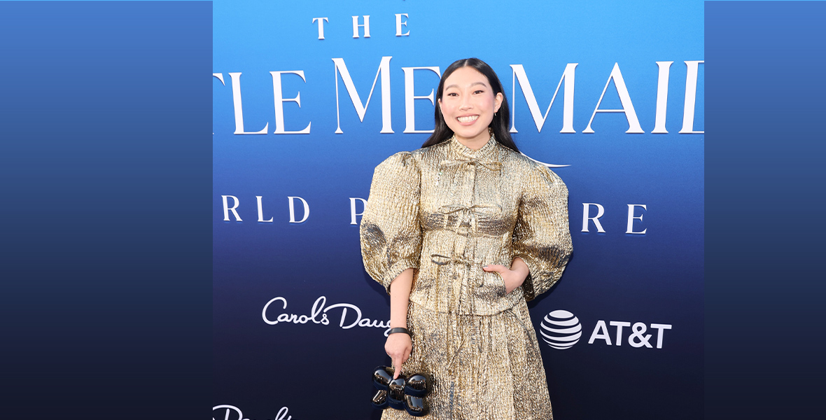 Awkwafina stands in front of the step and repeat for The Little Mermaid (2023), wearing a gold, sparkly dress and holding a small, black purse in her right hand.