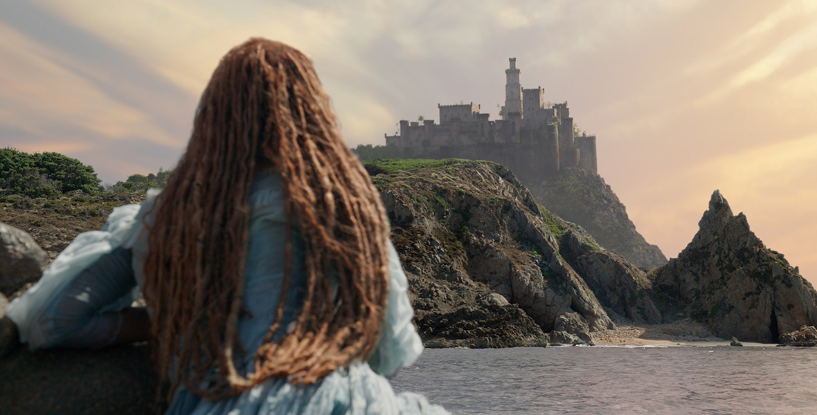 Ariel (Halle Bailey) sits with her back toward the camera, wearing a blue corseted dress, with her long red-hair flowing down her back, as she gazes at Prince Eric’s castle in the distance in a scene from The Little Mermaid.