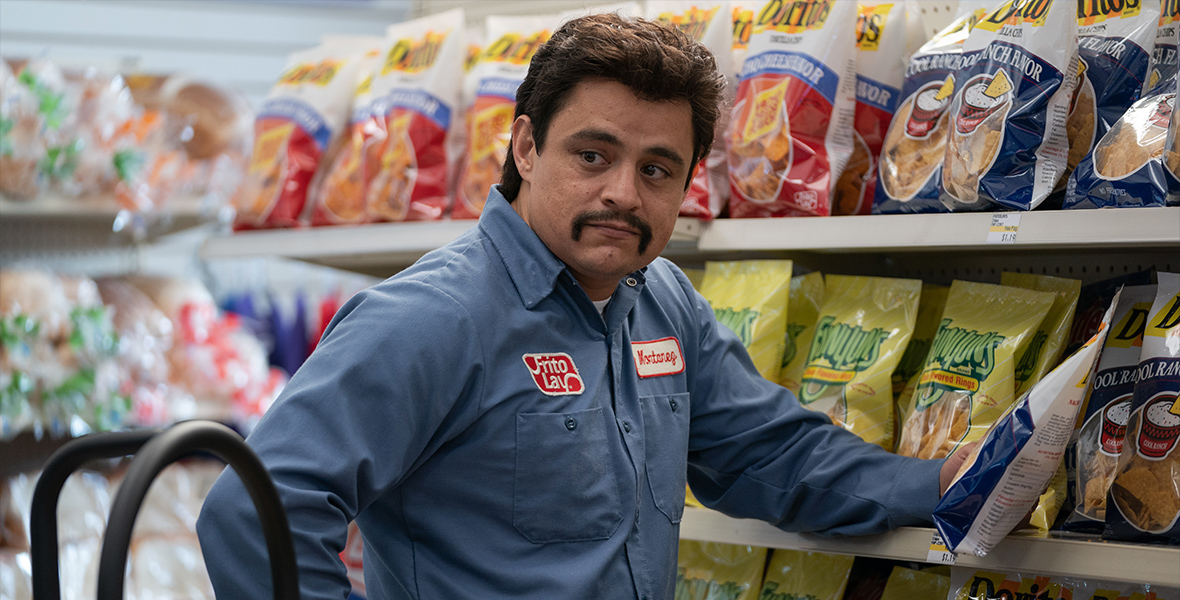 In a scene from Flamin’ Hot, Jesse Garcia, playing Richard Montañez, wears a Frito Lay uniform and stocks a grocery store shelf with bags of chips.