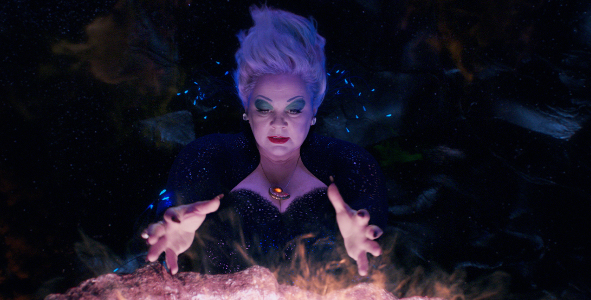 Ursula (Melissa McCarthy), using her magic, looks in on what Ariel has been up to on land. She stands facing the camera, with her hands posed over the scene she has conjured, wearing a sparkling gown, with the shell necklace holding Ariel’s voice within it.
