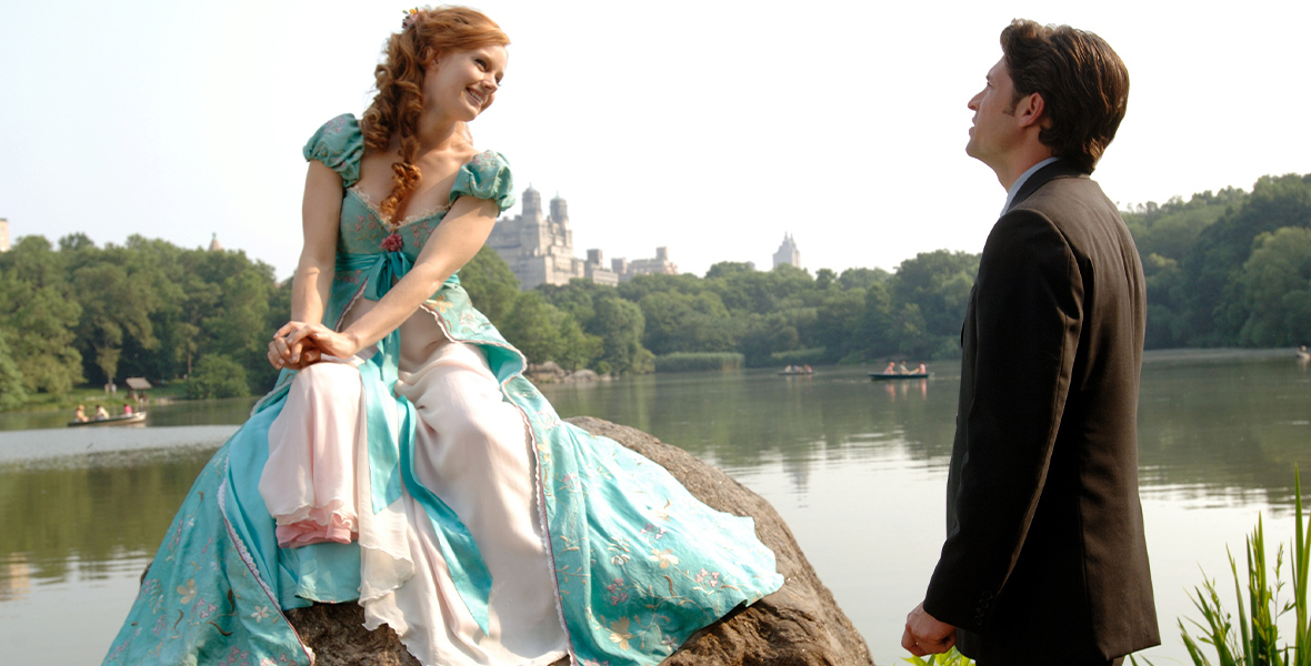 In Enchanted, Giselle sits atop a rock in Central Park. She wears a turquoise gown she crafted from curtains and smiles over at Robert. Robert wears a suit and looks confused by the musical number that just ended.