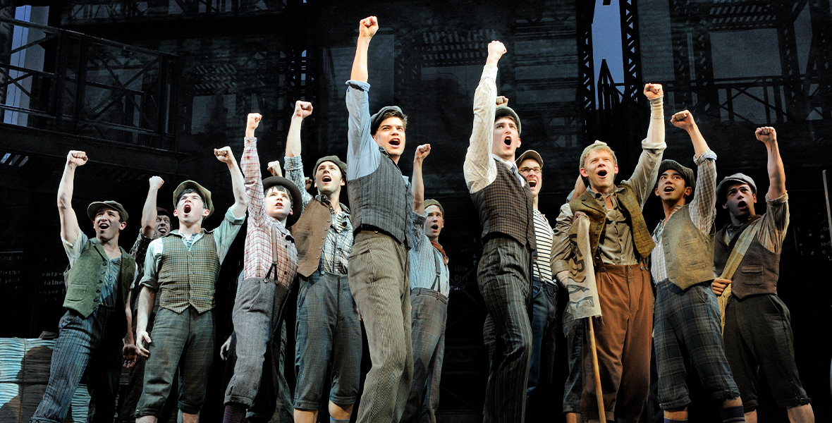 In Newsies: The Broadway Musical, Jack Kelly stands onstage with several “newsies” as they all raise their fists in the air. They wear newsboy caps and clothes reminiscent of the end of the 19th century.