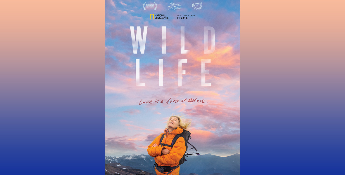 In the poster for National Geographic’s WILD LIFE, Kristine Tompkins is wearing an orange puffer jacket and a backpack, and is staring up at the sky; her hair is windswept. There are mountains behind her, and the sky is full of pink and white clouds. There are mountains beneath and behind her, and the logo for the film is seen above her.