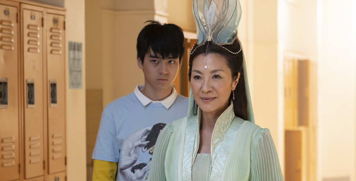 In an image from Disney+’s American Born Chinese, Wei-Chen (Jimmy Liu), in his highschooler form, is standing behind Guanyin (Michelle Yeoh), who is dressed in beautiful traditional-type garb in a light turquoise color. They’re standing in the high school hallway, and are both looking offscreen to the left.
