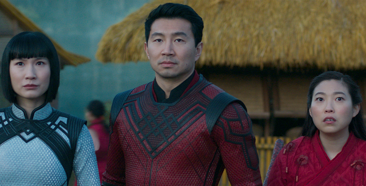 In a still from Marvel Studios’ Shang-Chi and The Legend of The Ten Rings, Xialing (Meng’er Zhang), Shang-Chi (Simu Liu) and Katy (Awkwafina) are wearing their fighting outfits and looking concerned. There are several thatched huts behind them.