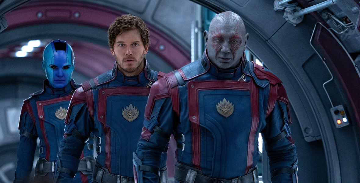 In a still from Marvel Studios' Guardians of the Galaxy Vol. 3, from left to right (and back to front,) Nebula (Karen Gillan), Peter Quill (Chris Pratt), and Drax (Dave Bautista) are walking down a spaceship hallway wearing matching uniforms.