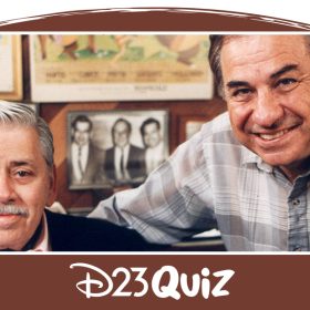 A color photo of Robert and Richard Sherman sitting in front of various posters from their projects. Robert has silvery white hair and a mustache. He’s wearing a light pink button-up shirt, black suit jacket, and black ascot. Richard’s hair is dark brown and he is wearing a gray plaid button-up and has a black wristwatch on his left hand. Behind the brothers you can make out posters for The Jungle Book, Mary Poppins, and Tom Sawyer.