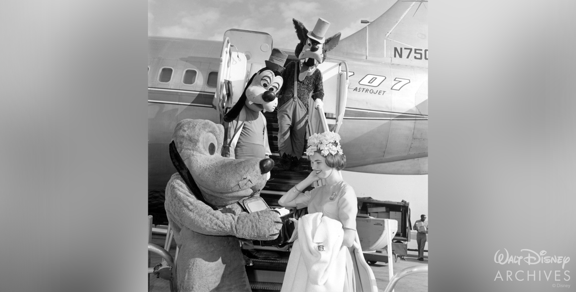 Standing at the bottom of the airstair of a passenger jet, Valerie Curry, wearing a dress, donning flowers in her hair, and holding a coat, is greeted by Pluto. Standing on the stairs closer to the jet’s entry door are Goofy and the Big Bad Wolf, who look down at Pluto and Curry.