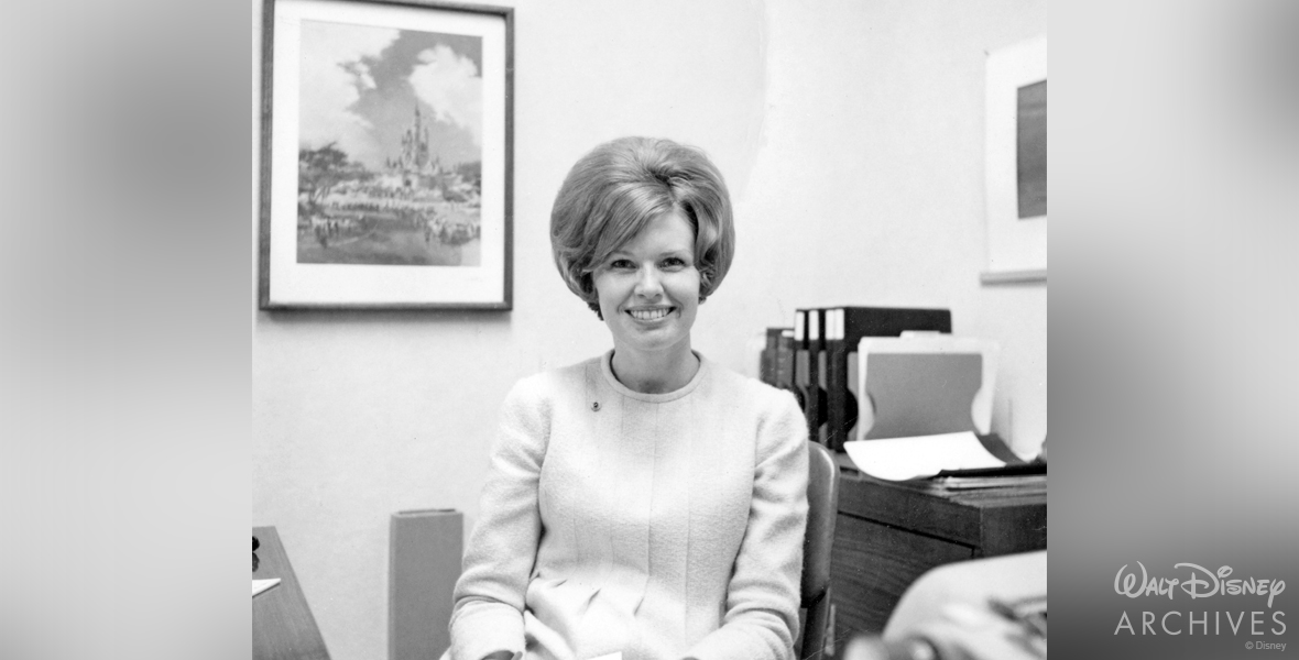 Valerie Curry is seated at an office desk at WED Enterprises in California. Behind her is framed concept art of Cinderella Castle from Walt Disney World. Also behind Curry, to her left, are file folders and other office materials.