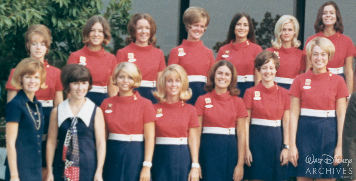 Valerie Curry stands with thirteen fellow Cast Members outside the Walt Disney World Preview Center in Lake Buena Vista, Florida. The Cast Members are arranged in two rows, one in front of the other, with seven in each row. Curry is positioned in the front row, on the far left, and behind her is green foliage.