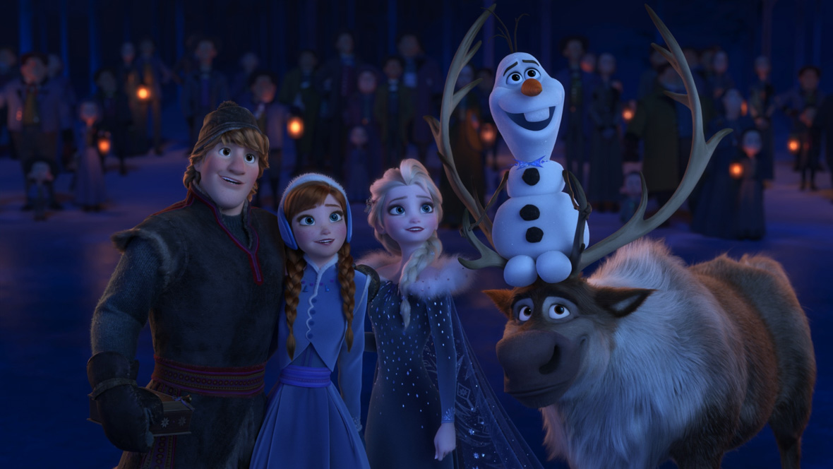 In this image from Olaf’s Frozen Adventure, from left to right, Kristoff (voiced by Disney Legend Jonathan Groff), Anna (voiced by Disney Legend Kristen Bell), Elsa (voiced by Disney Legend Idina Menzel), Olaf (voiced by Disney Legend Josh Gad), and reindeer Sven are standing together and looking at something in the sky to the right. They have smiles on their faces. A blurry crowd is seen behind them, against a snowy landscape; some are holding lanterns. Everyone is wearing coats and mittens.