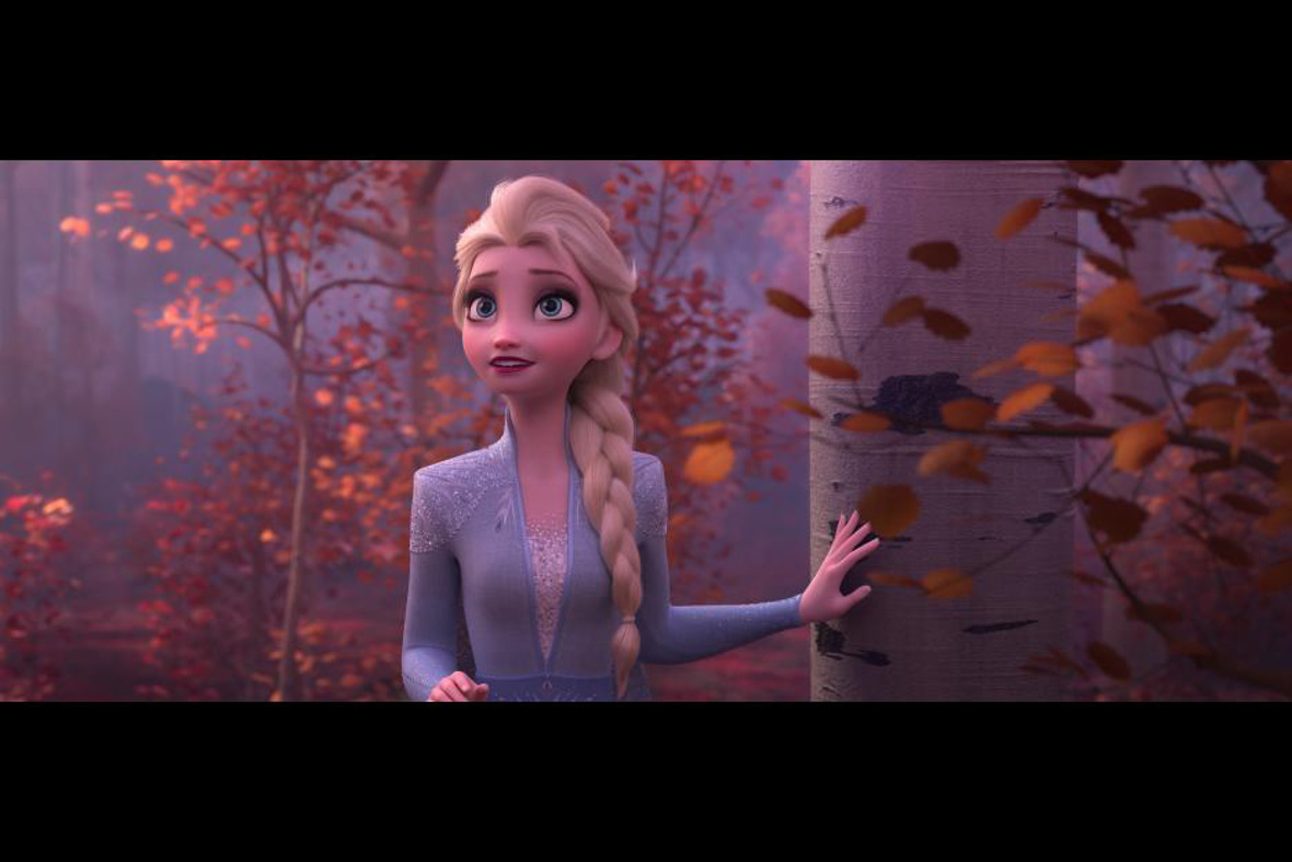In an image from Frozen 2, Elsa (Disney Legend Idina Menzel) is seen standing in a forest, among trees with light-colored trunks and small red and orange leaves. She has one hand one a trunk and is looking off at something in the sky to the left. She’s wearing a light blue tunic and her blonde hair is in a braid.