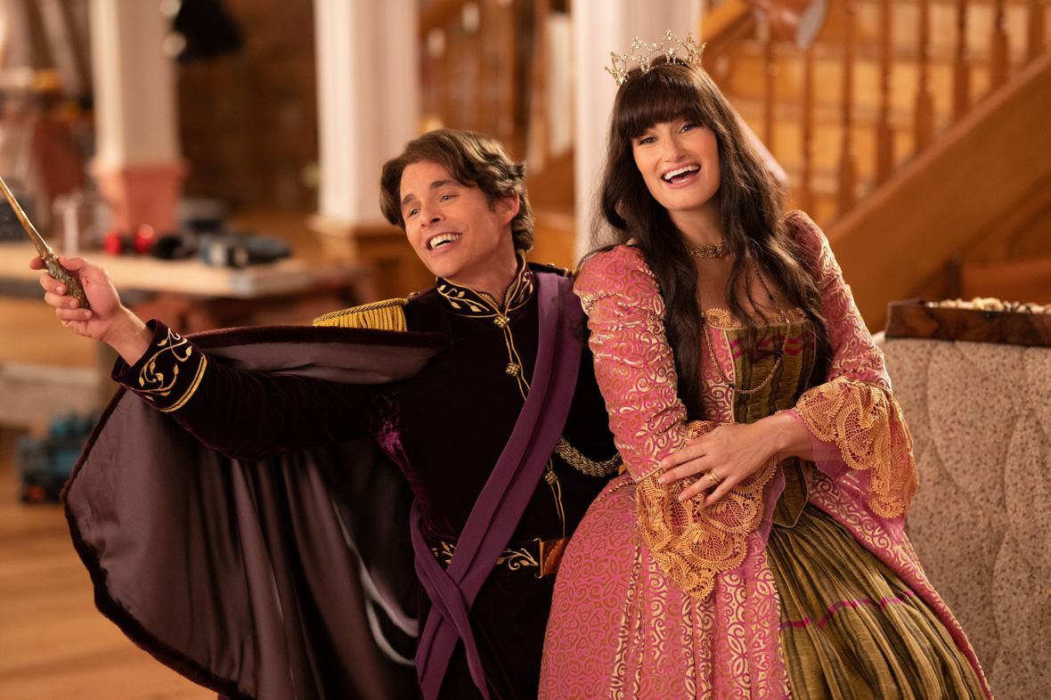 In an image from Disenchanted, Nancy (Disney Legend Idina Menzel) is sitting on the knee of Prince Edward (James Marsden); they are both singing, and Edward has one arm outstretched, and he’s holding a wand. He is wearing a dark purple tunic and cape; she is wearing a pink and gold brocade gown and a thin gold crown. They’re in a living room of some kind—a couch and a stairway can be seen behind them.