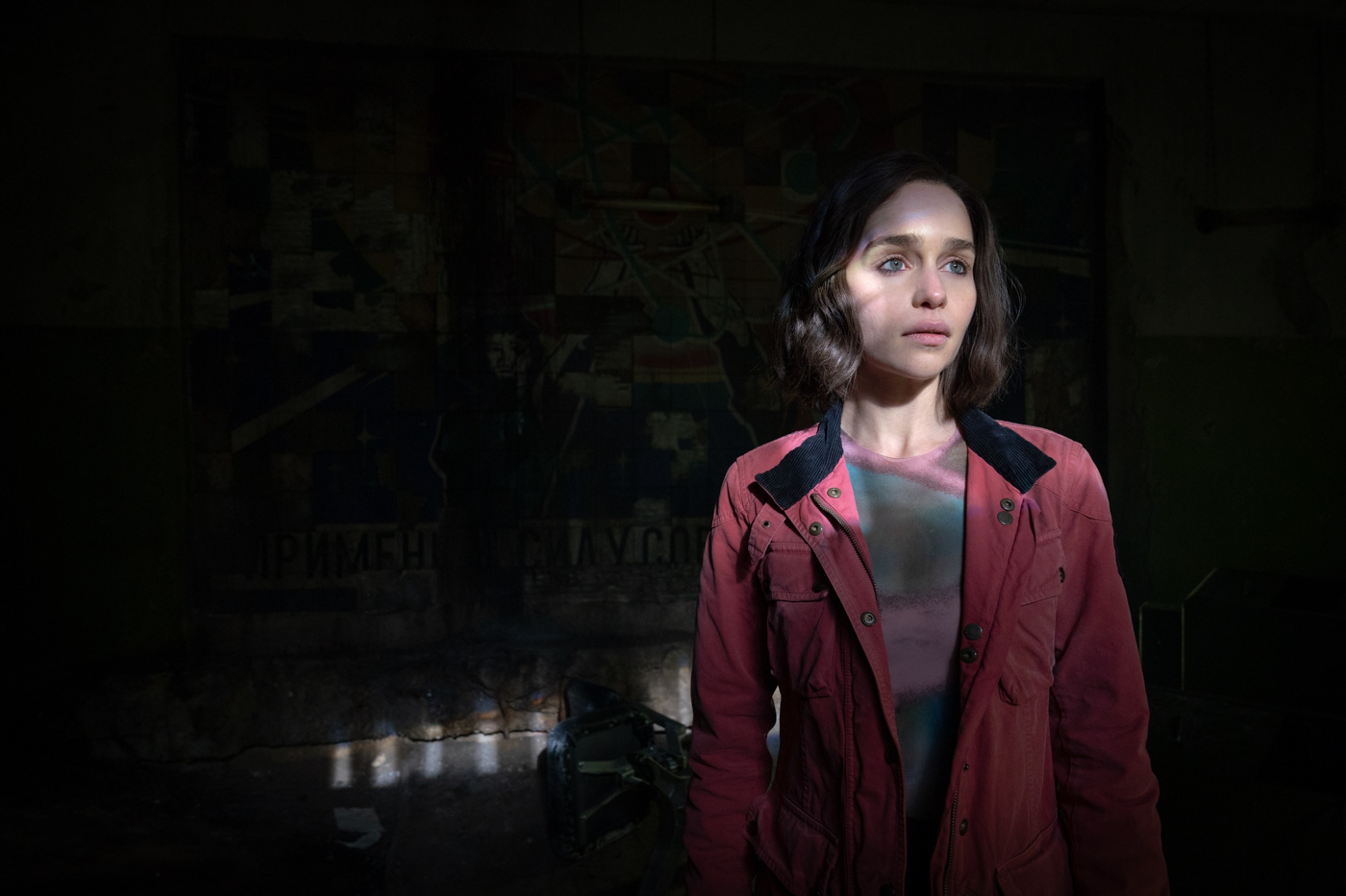 In a scene from the Marvel Studios series Secret Invasion, G’iah, played by Emilia Clarke, is standing in a dark cave with her face illuminated. She is wearing a pink and gray striped shirt and a deep pink jacket.