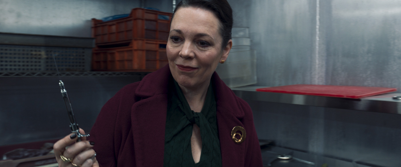 In a scene from the Marvel Studios series Secret Invasion, Sonya Falsworth, played by Olivia Colman, is holding a syringe with a needle attached to it. She is wearing a burgundy coat and black shirt.  