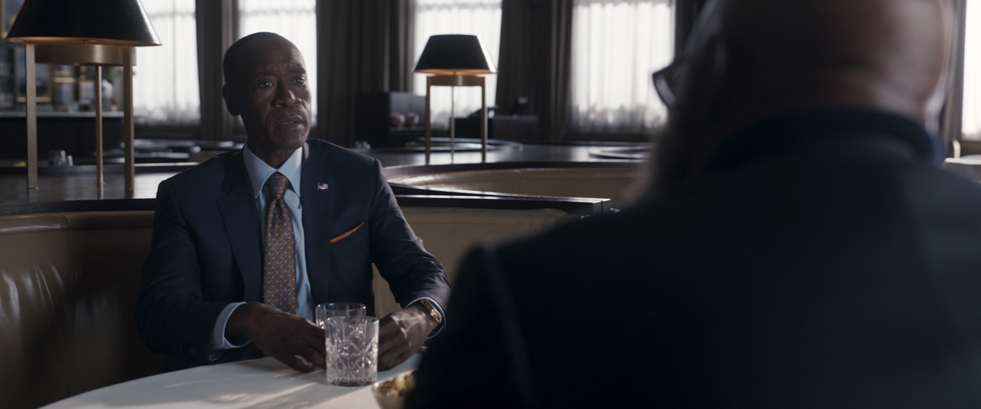 In a scene from the Marvel Studios series Secret Invasion, Colonel James Rhodes, played by Don Cheadle, is sitting across a table from Fury and wearing a blue blazer, maroon tie, and blue shirt while holding a glass.