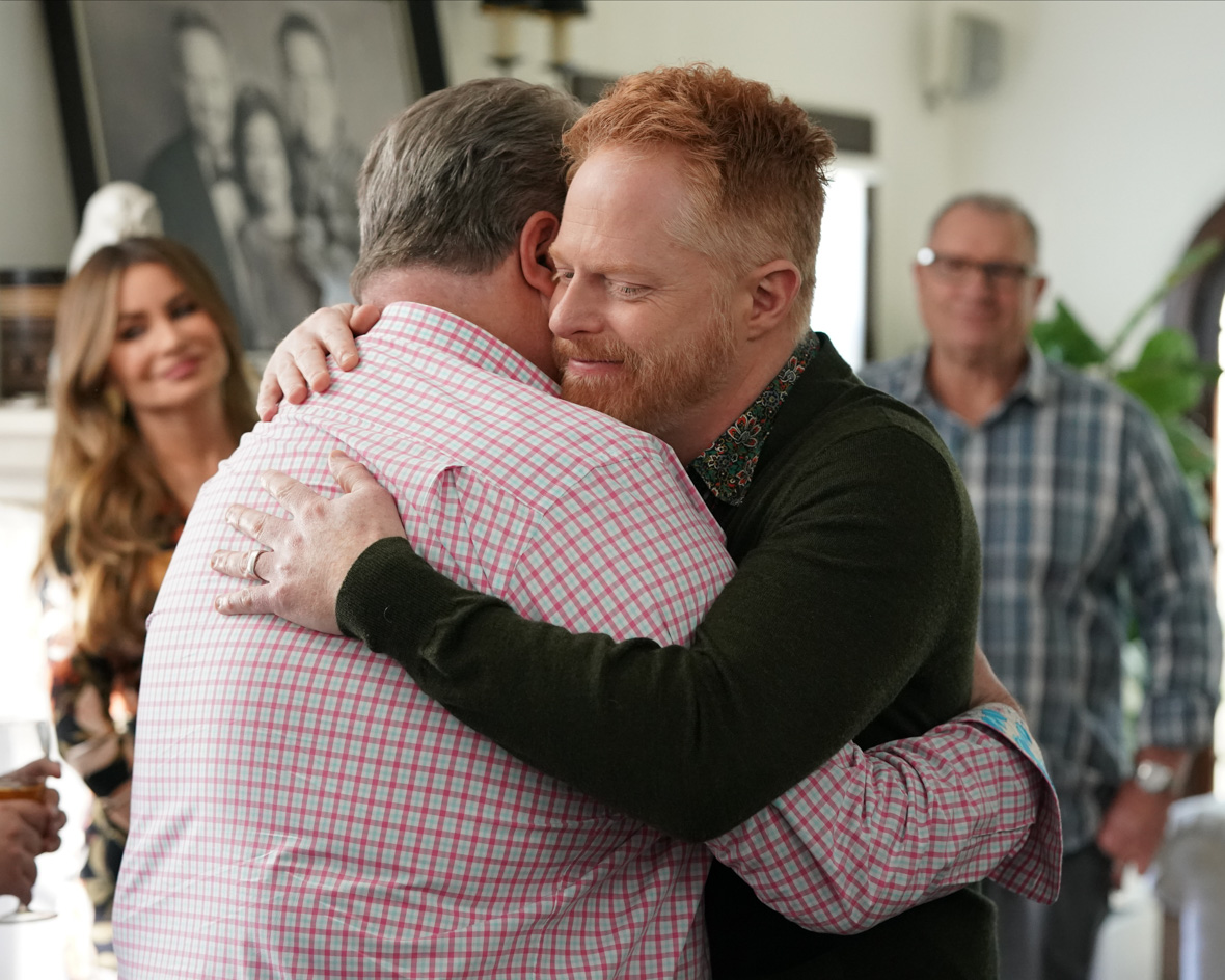 In the series Modern Family, husbands Mitch and Cam embrace in front of their family. Mitch has red hair and wears a green sweater, while Cam wears a long-sleeve red and white button-up. They stand inside their home with a portrait of their family behind them.