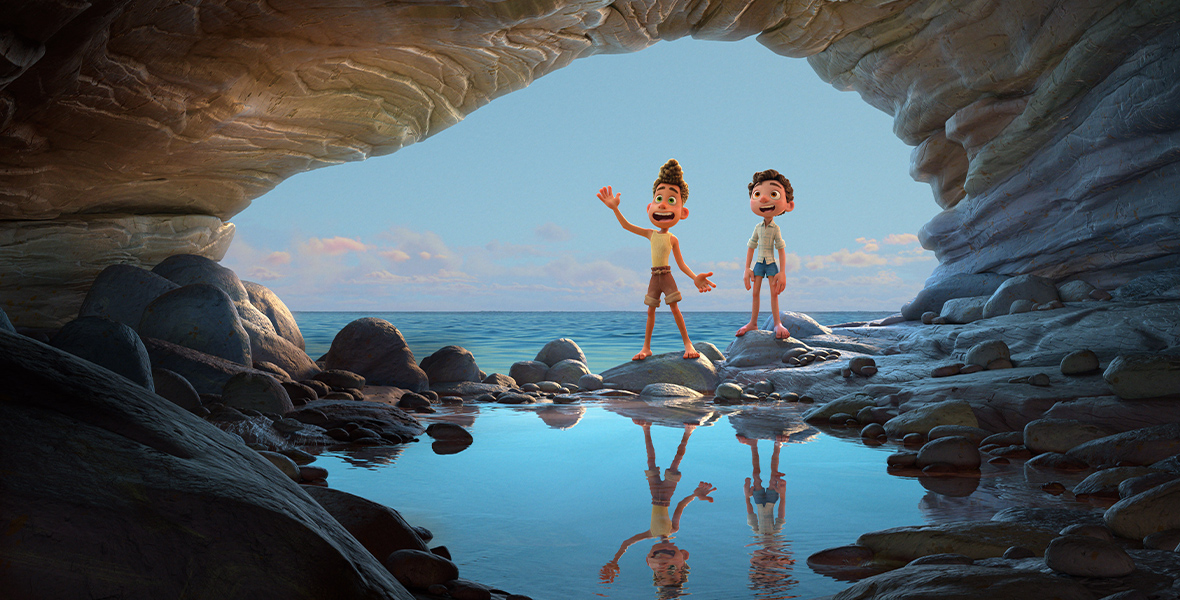 In the animated film Luca, two young boys—Luca and Alberto—stand on rocks at the mouth of a seaside cave. The ocean and bright blue sky are behind them, and there is a small pool inside the cave that shows their reflections. They grin.