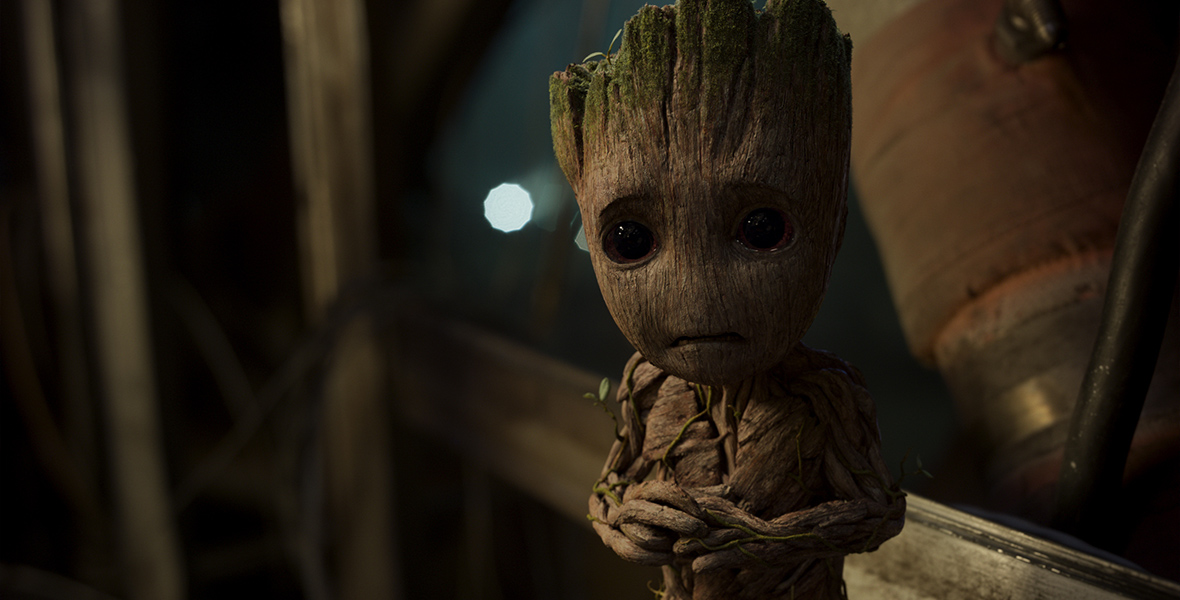 A still of baby Groot in Guardians of the Galaxy Vol 2. He is a tiny, tree-like creature with small vines growing around his bark and big brown eyes. He clasps his hands together and looks onward nervously.