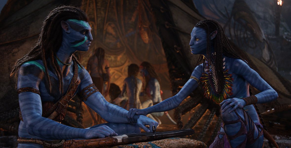 In an image from Avatar: The Way of Water, Jake Sully (Sam Worthington) and Neytiri (Zoe Saldaña) are facing each other and talking. Behind them are the rest of their family, inside a tent-type structure. It appears to be nighttime, and Jake and Neytiri’s conversation appears serious; Neytiri is grabbing Jake’s arm, and there is a firearm resting on a rock in front of him.