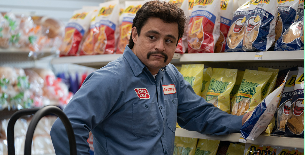 In an image from Flamin’ Hot, Richard Montañez (Jesse Garcia) stands next to a shelving unit full of bags of Frito Lay chips inside a grocery store. There is a hand trolley near him, and he’s wearing a shirt with a Frito Lay logo patch on it.