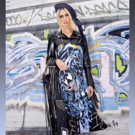 Hila Klein stands against a heavily graffitied wall wearing the Venom slip dress from her Marvel x TF collection. She has a navy-blue hat on top of straight blonde hair. Over the slip dress she has on a heavily cropped black leather jacket, so the full image of Venom is visible on her dress. Draped over one arm is a black fringe bag with a metal studded strap. She is gazing intensely at the camera and has a smokey eye, plus some white jewel studs across her eyes.