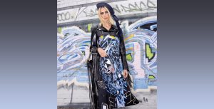 Hila Klein stands against a heavily graffitied wall wearing the Venom slip dress from her Marvel x TF collection. She has a navy-blue hat on top of straight blonde hair. Over the slip dress she has on a heavily cropped black leather jacket, so the full image of Venom is visible on her dress. Draped over one arm is a black fringe bag with a metal studded strap. She is gazing intensely at the camera and has a smokey eye, plus some white jewel studs across her eyes.
