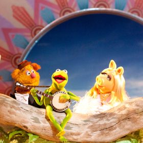 Kermit the Frog sits on a log, singing “The Rainbow Connection” and playing the banjo. Behind him he is joined by Scooter, the Swedish Chef, Fozzie, Miss Piggy, Sam the Eagle, and Beauregard. The stage behind them is decorated with a backdrop of the sky and a colorful border.