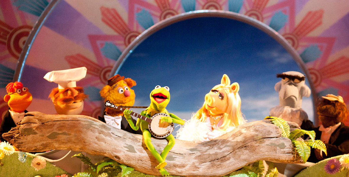 Kermit the Frog sits on a log, singing “The Rainbow Connection” and playing the banjo. Behind him he is joined by Scooter, the Swedish Chef, Fozzie, Miss Piggy, Sam the Eagle, and Beauregard. The stage behind them is decorated with a backdrop of the sky and a colorful border.