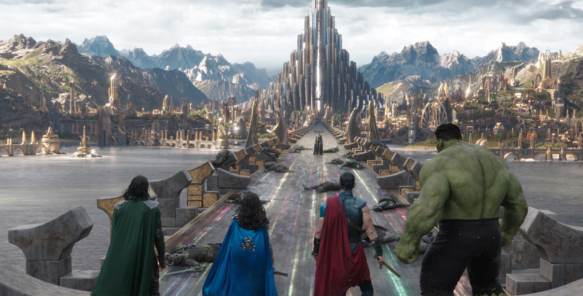 Loki, Valkyrie, Thor, and The Hulk stand on the Rainbow Bridge, facing the goddess Hela. The Rainbow Bridge leads into the city of Asgard, which sits among green mountaintops.