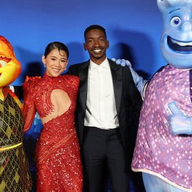 At the premiere of Elemental, Leah Lewis and Mamoudou Athie pose with the characters the voice, fiery Ember and water-based Wade. They all stand in front of a backdrop of Element City.