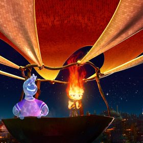 In this image from Disney and Pixar’s Elemental, Wade, a Water person (voiced by Mamoudou Athey), on the left, and Ember, a Fire person (voiced by Leah Lewis), on the right, stand in what appears to be a giant wok that is jury-rigged to a fabric hot air balloon that stretches off of the top of the frame. Ember’s fire is burning bright at the top of her head, heating the air keeping them afloat. The night sky is behind them, and below them are the fire-lit buildings of Fire Town.
