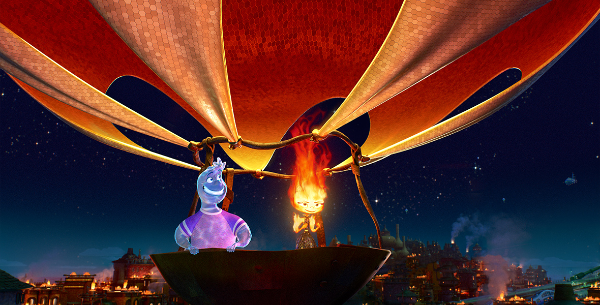In this image from Disney and Pixar’s Elemental, Wade, a Water person (voiced by Mamoudou Athey), on the left, and Ember, a Fire person (voiced by Leah Lewis), on the right, stand in what appears to be a giant wok that is jury-rigged to a fabric hot air balloon that stretches off of the top of the frame. Ember’s fire is burning bright at the top of her head, heating the air keeping them afloat. The night sky is behind them, and below them are the fire-lit buildings of Fire Town.