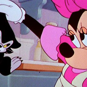 In this image from the animated short Bath Day, Minnie Mouse, on the right, holds up a dripping Figaro the cat with her gloved right hand. Figaro is frowning. Minnie is wearing a pink ribbon on her head and matching pink dress, partly covered by a white apron. The tiled wall and shelf of a bathroom is behind them.