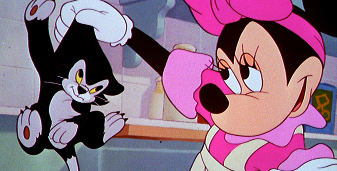 In this image from the animated short Bath Day, Minnie Mouse, on the right, holds up a dripping Figaro the cat with her gloved right hand. Figaro is frowning. Minnie is wearing a pink ribbon on her head and matching pink dress, partly covered by a white apron. The tiled wall and shelf of a bathroom is behind them.