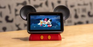 The Hey Disney device sits on a coffee table. The device is a tablet with mouse ears, stylized to look like Mickey Mouse, featuring an image of Donald, Minnie, and Mickey on the screen.
