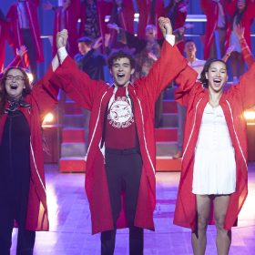 In a scene from Season 4 of High School Musical: The Musical: The Series, Carlos (Frankie Rodriguez), Ashlyn (Julia Lester), Ricky (Joshua Bassett), Gina (Sofia Wylie), and Kourtney (Dara Reneé) wear red graduation gowns and hold raised hands in the air. They are performing in the Senior Year Spring Musicale at East High alongside other classmates.