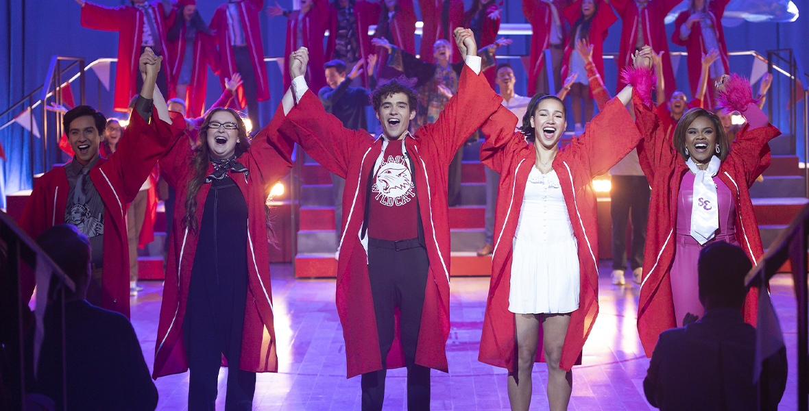 In a scene from Season 4 of High School Musical: The Musical: The Series, Carlos (Frankie Rodriguez), Ashlyn (Julia Lester), Ricky (Joshua Bassett), Gina (Sofia Wylie), and Kourtney (Dara Reneé) wear red graduation gowns and hold raised hands in the air. They are performing in the Senior Year Spring Musicale at East High alongside other classmates.
