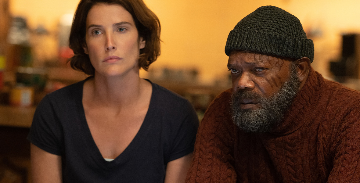 In an image from Marvel Studios’ Secret Invasion, from left to right, Maria Hill (Cobie Smulders) and Nick Fury (Samuel L. Jackson) sit next to each other inside a warmly lit room. They have concerned looks on their faces. Maria is wearing a dark V-neck T-shirt; Nick is wearing a dark red sweater turtleneck and a black beanie; he is not wearing his usual eyepatch.