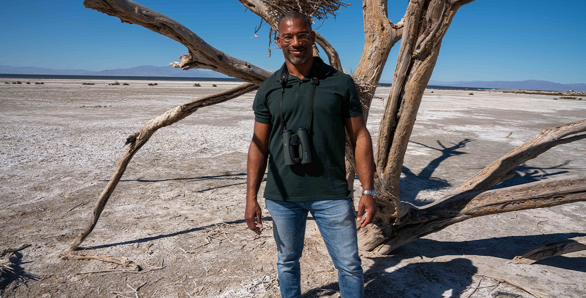 In an image from National Geographic’s Extraordinary Birder with Christian Cooper, host Christian Cooper is standing in the Salton Sea, a vital stop for migrating birds. Behind him is a dead tree with nests that used to be homes to Great Blue Herons.