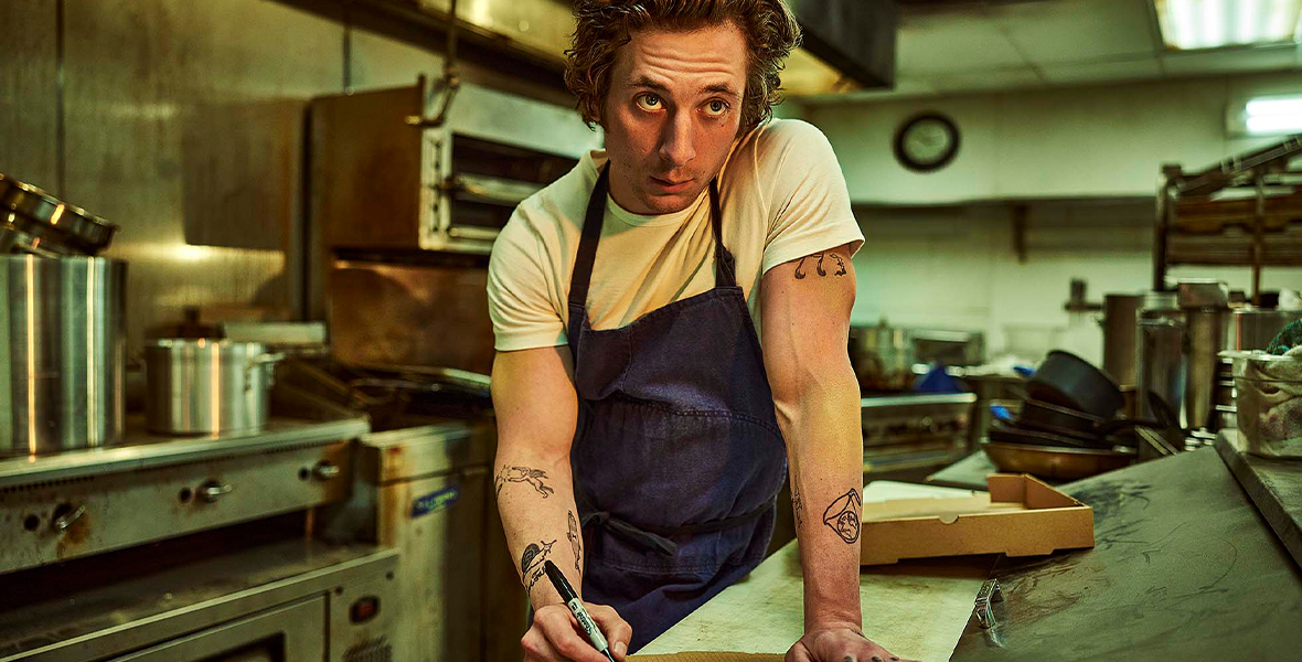 In an image from FX’s The Bear, Carmen “Carmy” Berzatto (Jeremy Allen White) is standing in his restaurant’s kitchen. He’s leaning over a counter and writing out a menu on the top of a pizza box. His arms are covered with tattoos and he wears an apron. Behind him are various ovens and burners, with pots and pans strewn about.