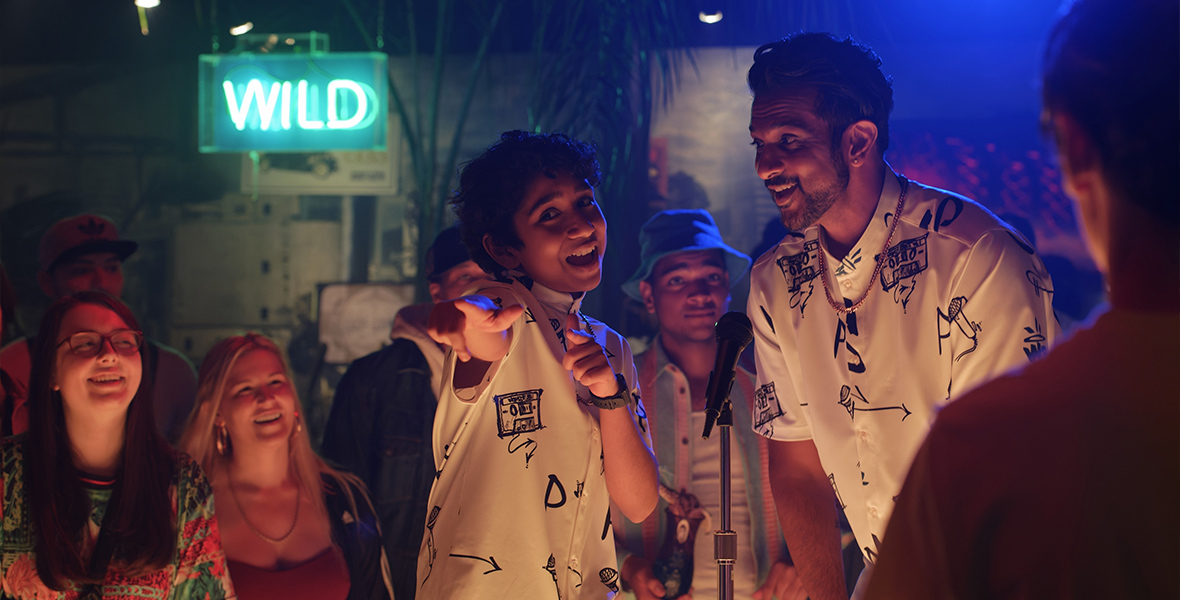 In an image from the Disney+ original movie World’s Best, Prem (Manny Magnus) is seen performing with his father (Utkarsh Ambudkar); they’re both wearing black and white graphic button-down shirts, and are standing behind a microphone on a mic stand. The room is relatively dark, but you can see a crowd of people surrounding them, and light is shining on everyone’s faces from below. A neon sign that says “Wild” is seen on the wall behind them.