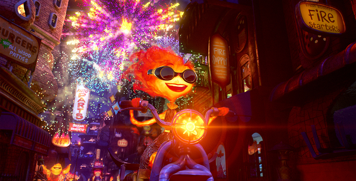 In this image from Disney and Pixar’s Elemental, Ember, a Fire person (voiced by Leah Lewis), pilots a scooter through the streets of Fire Town, which has an orange headlight and is coming toward the camera. Ember is smiling and wearing dark goggles. Fireworks light up the street behind her.