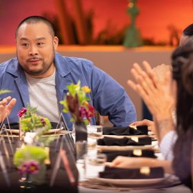 In an image from the new Hulu competition series Secret Chef, executive producer and chef David Chang is sitting at the head of a table, wearing a blue long-sleeved shirt with a white T-shirt underneath. Several of the show’s contestants are also sitting at the table, seen to his right—and there are plates, silverware, and flowers in vases in front of them; they are facing Chang as he speaks.